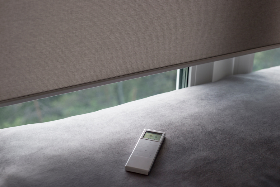 White remote control panel for motorized roller shades. Automatic roller blinds beige color on the window. Remote Control Shades are near the sofa. Remote control lies on the sofa.